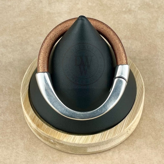The bracelet features a matte metallic copper leather band, elegantly curved with a long, arcing, antique silver magnetic clasp. Displayed on a black conical stand with a natural wood base, it blends warm tones with sleek silver. The ergonomic clasp ensures a secure and comfortable fit, while the rich leather texture contrasts beautifully with the smooth silver for a bold yet timeless look.