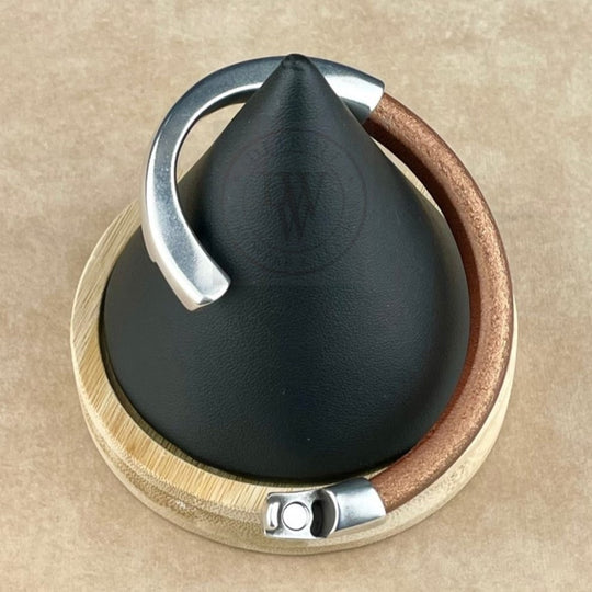 The bracelet features a matte metallic copper leather band, elegantly curved with a long, arcing, antique silver magnetic clasp. Displayed on a black conical stand with a natural wood base, it blends warm tones with sleek silver. The ergonomic clasp ensures a secure and comfortable fit, while the rich leather texture contrasts beautifully with the smooth silver for a bold yet timeless look.