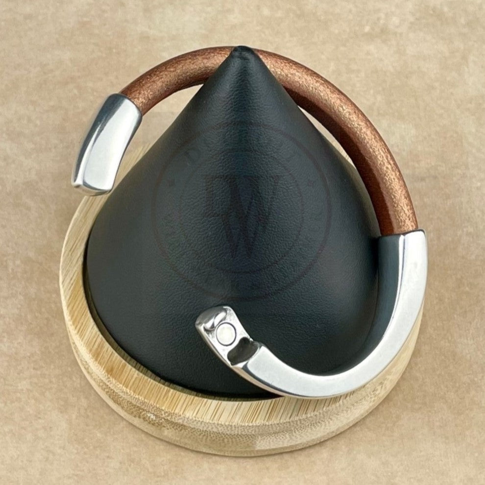 The bracelet features a matte metallic copper leather band, elegantly curved with a long, arcing, antique silver magnetic clasp. Displayed on a black conical stand with a natural wood base, it blends warm tones with sleek silver. The ergonomic clasp ensures a secure and comfortable fit, while the rich leather texture contrasts beautifully with the smooth silver for a bold yet timeless look.