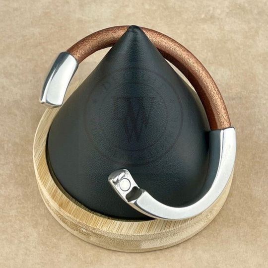 The bracelet features a matte metallic copper leather band, elegantly curved with a long, arcing, antique silver magnetic clasp. Displayed on a black conical stand with a natural wood base, it blends warm tones with sleek silver. The ergonomic clasp ensures a secure and comfortable fit, while the rich leather texture contrasts beautifully with the smooth silver for a bold yet timeless look.