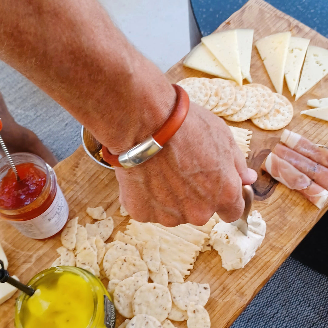 A person wearing an orange leather bracelet with silver clasp is preparing a cheese and cracker platter on a wooden board. Cobo bracelet lifestyle photo.