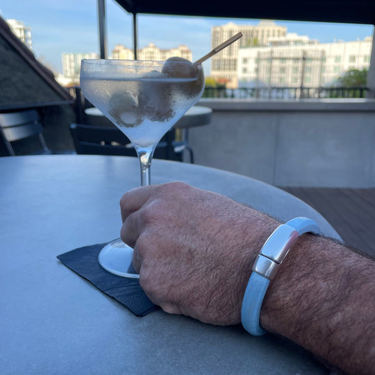 Hand holding a martini glass with olives and wearing a light blue leather Cobo bracelet on a rooftop with a cityscape view. Cobo bracelet lifestyle photo.