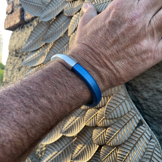 A close-up of a wrist wearing a blue Arena leather bracelet with a silver clasp on a textured surface. Arena bracelet lifestyle photo.