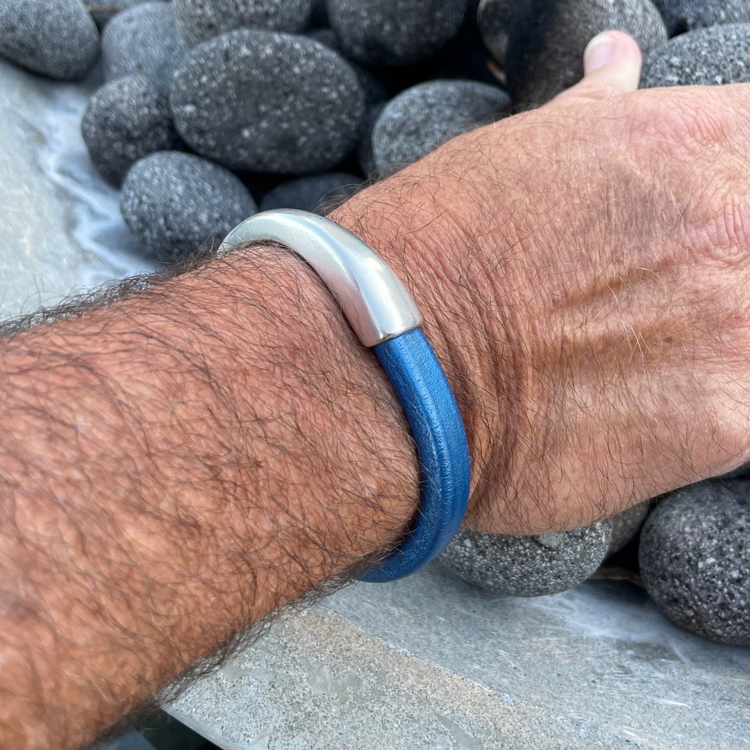 Hand wearing a blue Arena bracelet with a silver clasp against a background of gray stones. Arena bracelet lifestyle photo.