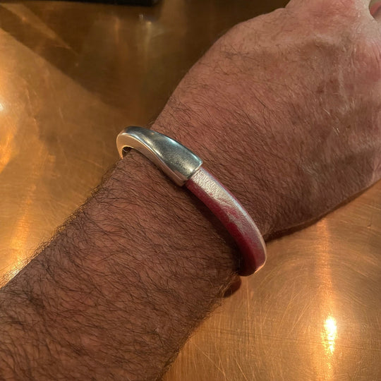 Close-up of a wrist wearing a soft silver-washed red leather Arena bracelet with a silver clasp on a reflective copper surface. Arena bracelet lifestyle photo.