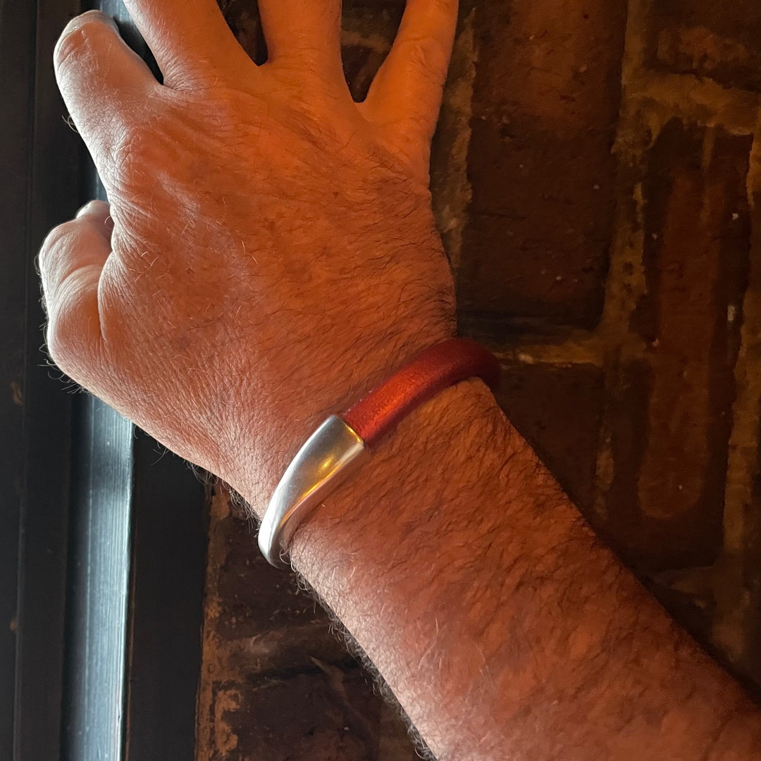 A close-up of a wrist wearing a rust colored leather bracelet with a silver clasp on a textured surface. Arena bracelet lifestyle photo.