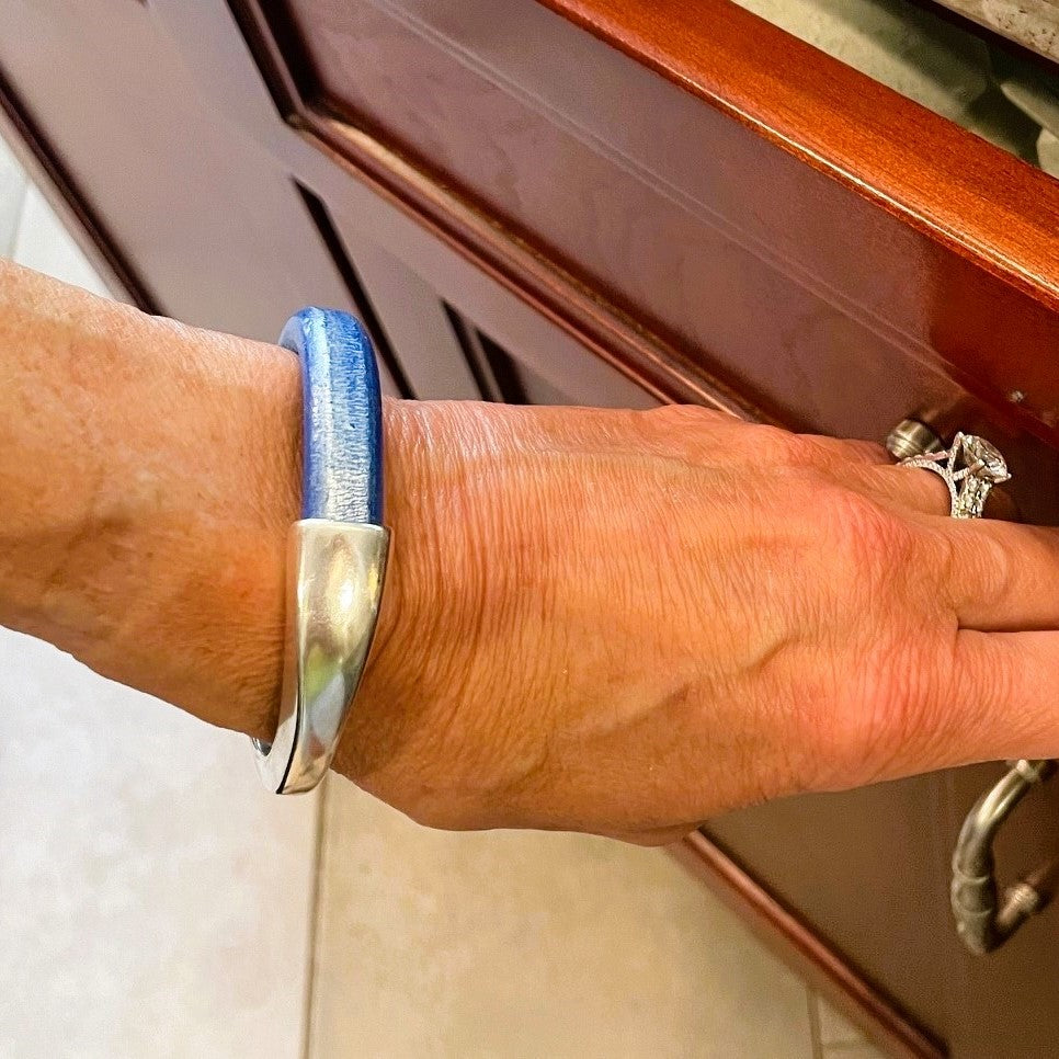 A hand wearing a blue leather Arena bracelet with a silver clasp and cabinets in the background. Arena bracelet lifestyle photo.