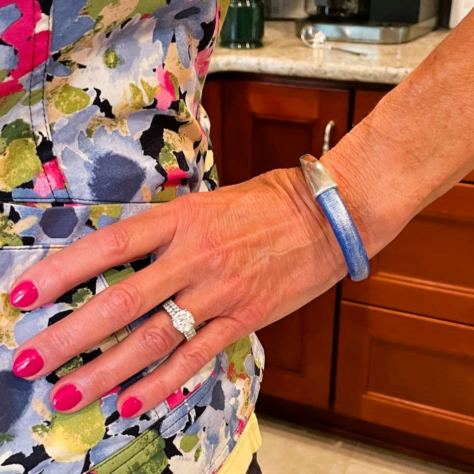 Close-up of a hand with pink nail polish wearing a diamond ring and a blue leather Arena bracelet, with a colorful floral-patterned garment and wooden cabinets in the background. Arena bracelet lifestyle photo.