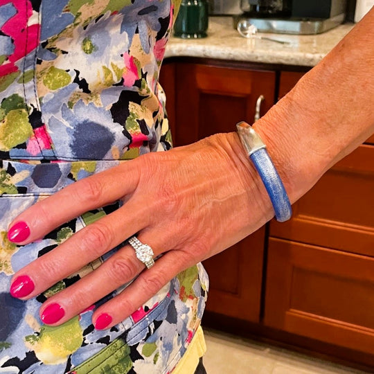 Close-up of a hand with pink nail polish wearing a diamond ring and a blue leather Arena bracelet, with a colorful floral-patterned garment and wooden cabinets in the background. Arena bracelet lifestyle photo.