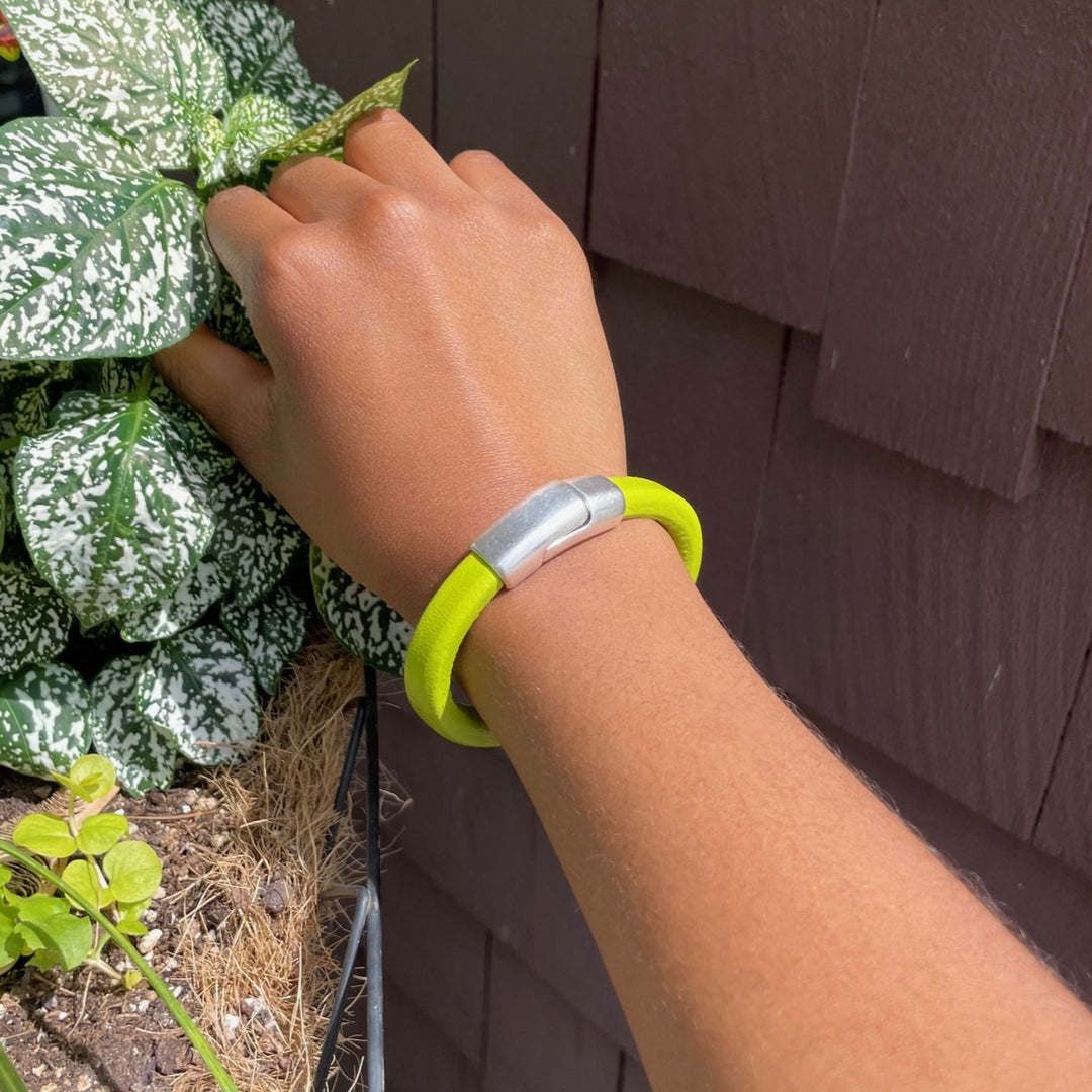 A wrist wearing a bright lime green Cobo leather bracelet, and pinching back a leafy plant against a wooden wall. Cobo bracelet lifestyle photo.
