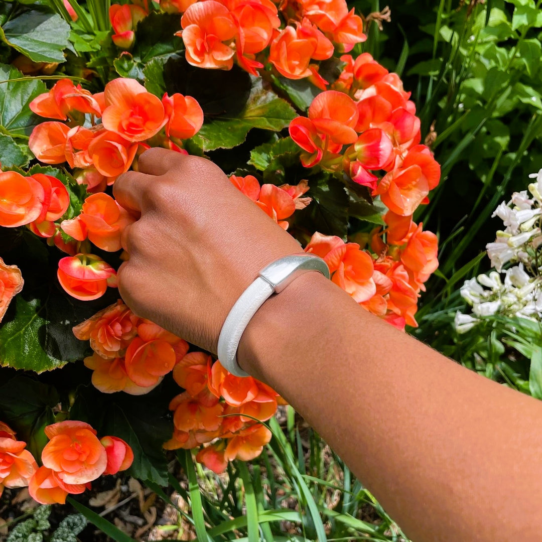 A hand reaching towards orange begonia flowers, and wearing a metallic pearl white Arena bracelet with a silver clasp. Arena bracelet lifestyle photo.
