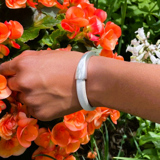 A hand reaching towards orange begonia flowers, and wearing a metallic pearl white Arena bracelet with a silver clasp. Arena bracelet lifestyle photo.