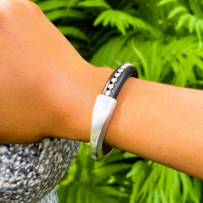 A clear crystal chain embedded in a black leather Arena bracelet with a silver clasp on a wrist with a blurred green background. Arena bracelet lifestyle photo.