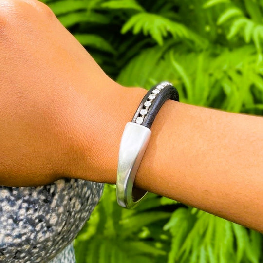 A clear crystal chain embedded in a black leather Arena bracelet with a silver clasp on a wrist with a blurred green background. Arena bracelet lifestyle photo.