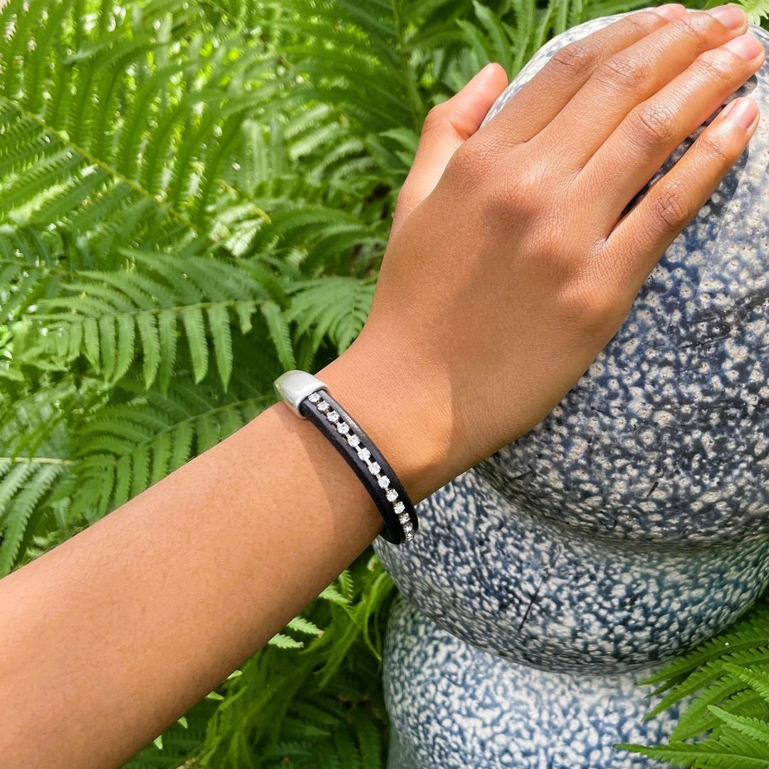A person wearing a crystal chain and black leather bracelet with green foliage and a blue and white sculpture in the background. Arena bracelet lifestyle photo. 