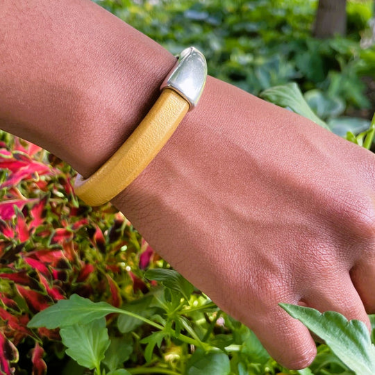 A wrist wearing a metallic gold Arena bracelet with a silver clasp against a green outdoor background. Arena bracelet lifestyle photo.