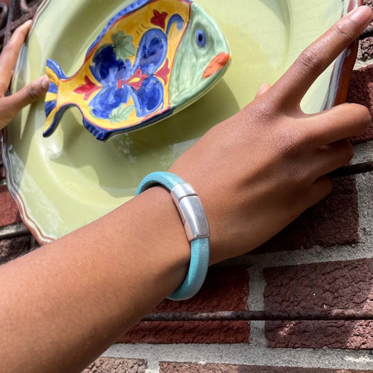 A close-up of a person wearing a blue leather Cobo bracelet while hanging a colorful ceramic fish on a green plate on a brick wall. Cobo bracelet lifestyle photo.