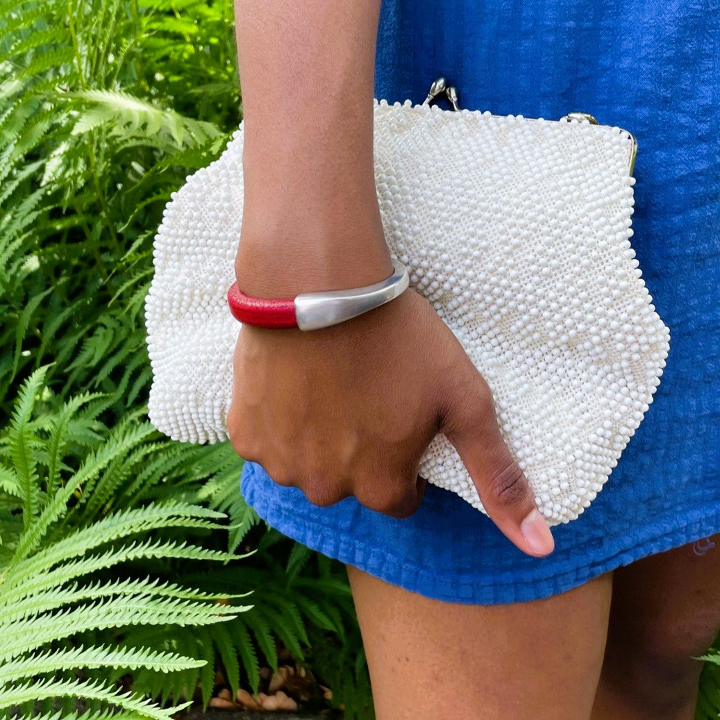 A person wearing a red Arena bracelet with a silver clasp and holding a white beaded clutch with a blue dress against a green fern background. Arena bracelet lifestyle photo.