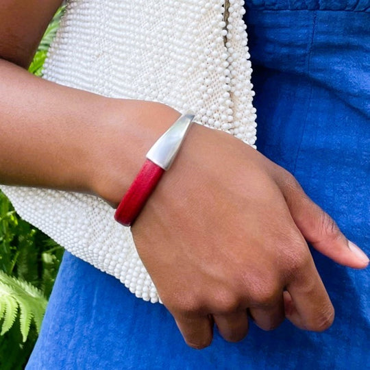 Close-up of a wrist wearing a red Arena bracelet with a silver clasp and holding a white beaded clutch with a blue dress against a green fern background. Arena bracelet lifestyle photo.