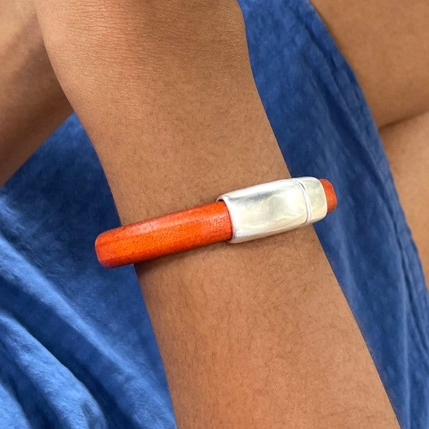 Close-up of an orange leather Cobo bracelet with a silver clasp on a wrist against a blue dress. Cobo bracelet lifestyle photo.