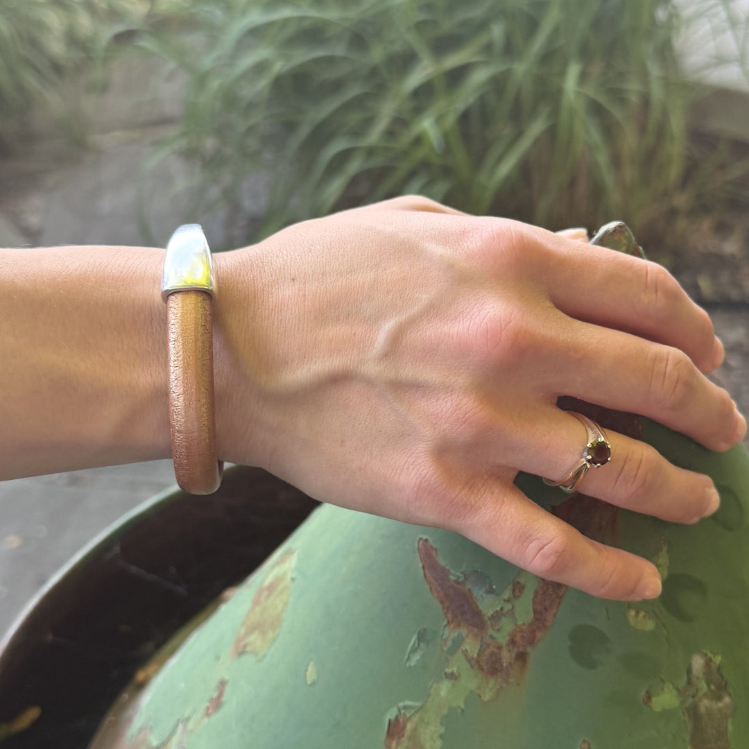 Hand wearing a brown leather bracelet and ring, holding a green object with a natural background